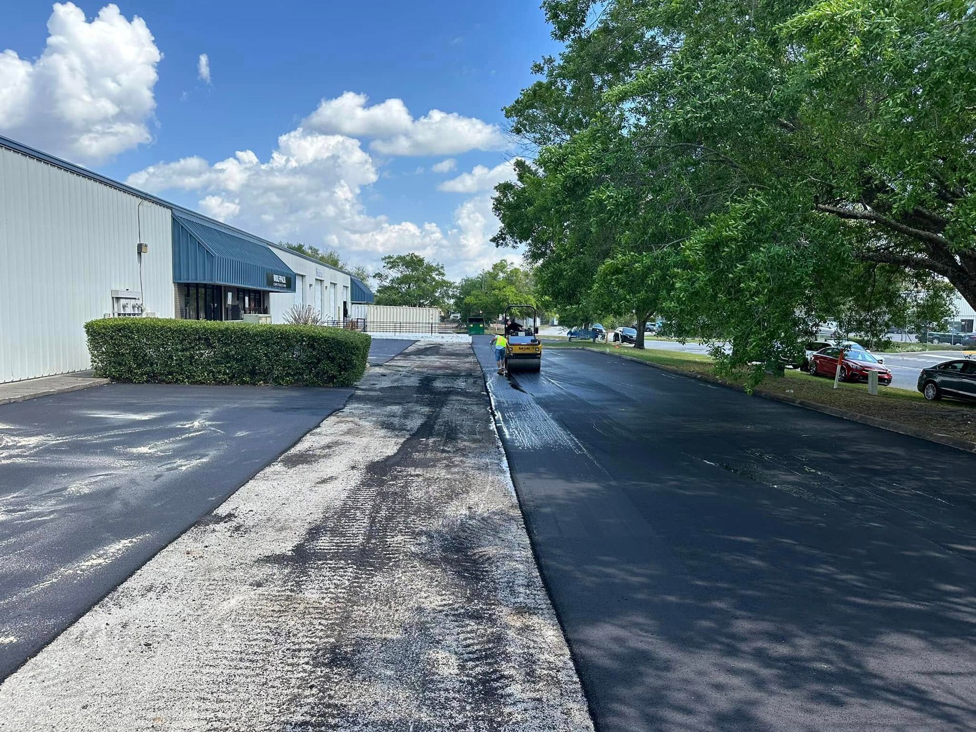 Industrial parking lot with a tree-lined driveway, parked cars, and a person riding a scooter under a blue sky