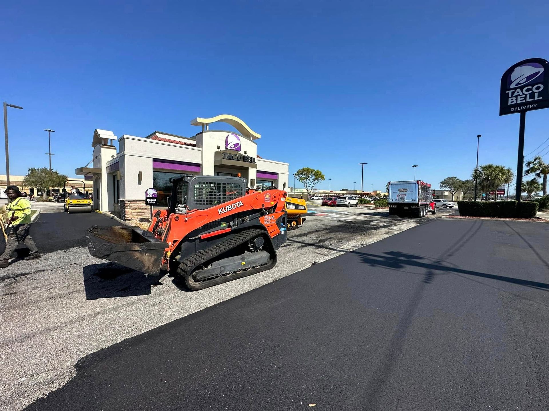 Orange skid steer on a road beside a building under construction, with fresh black pavement in front.