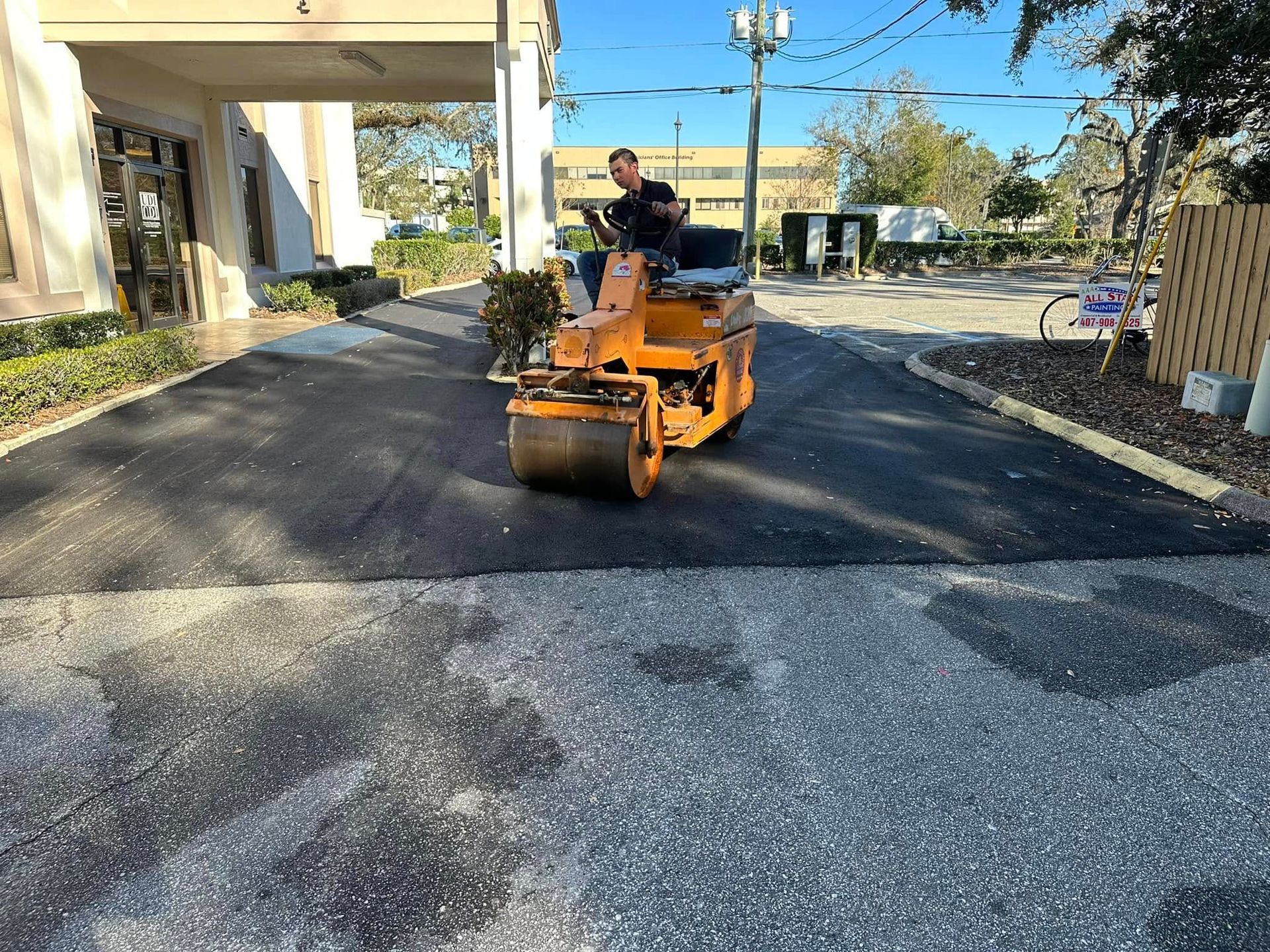 Orange road roller paving a dark asphalt driveway beside a building on a sunny day