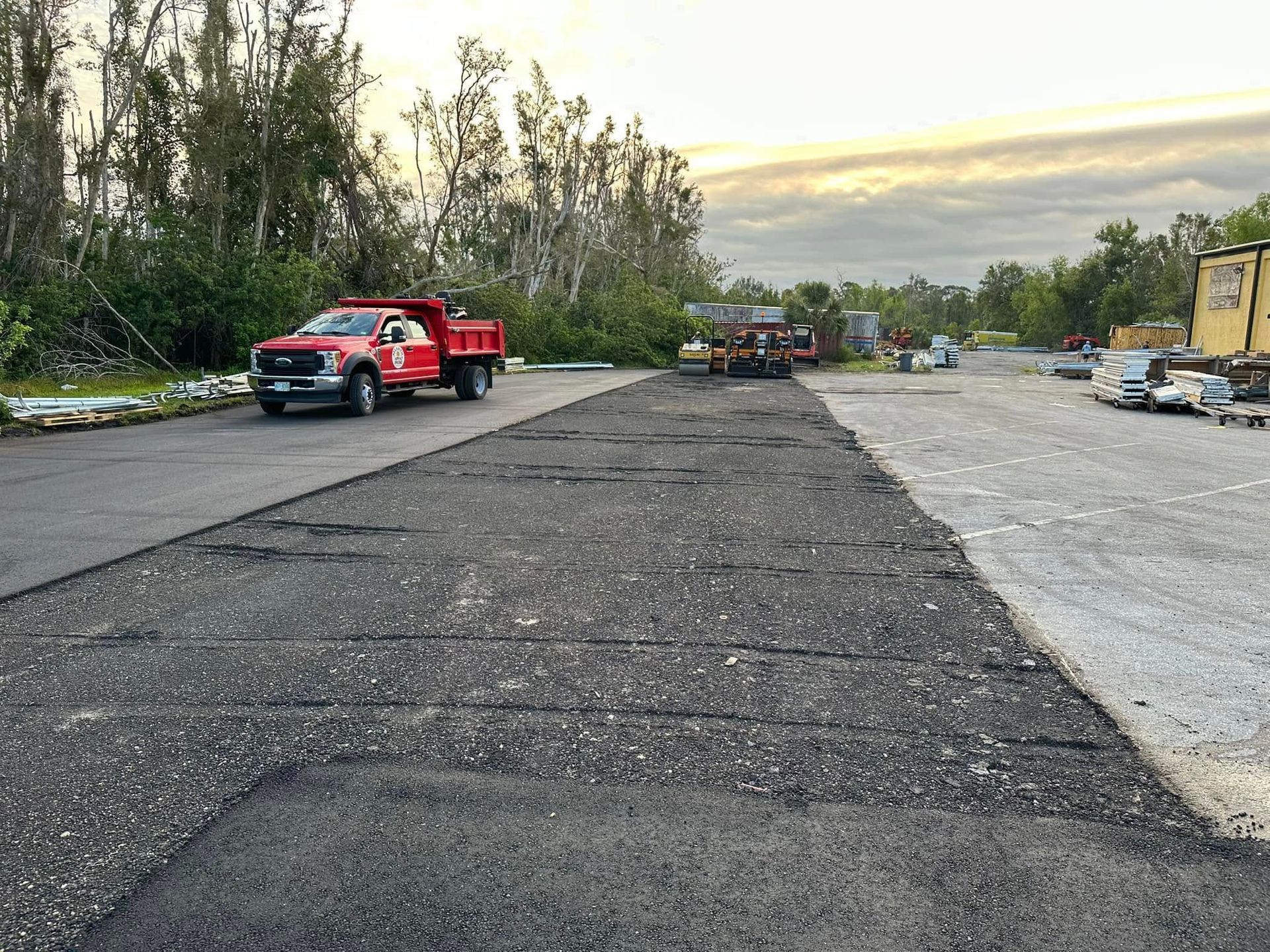 Red truck parked on a freshly paved lot beside gravel, with trees and construction equipment in the background