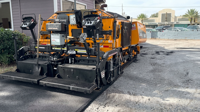 Orange construction machine paving gravel on a road beside a building.