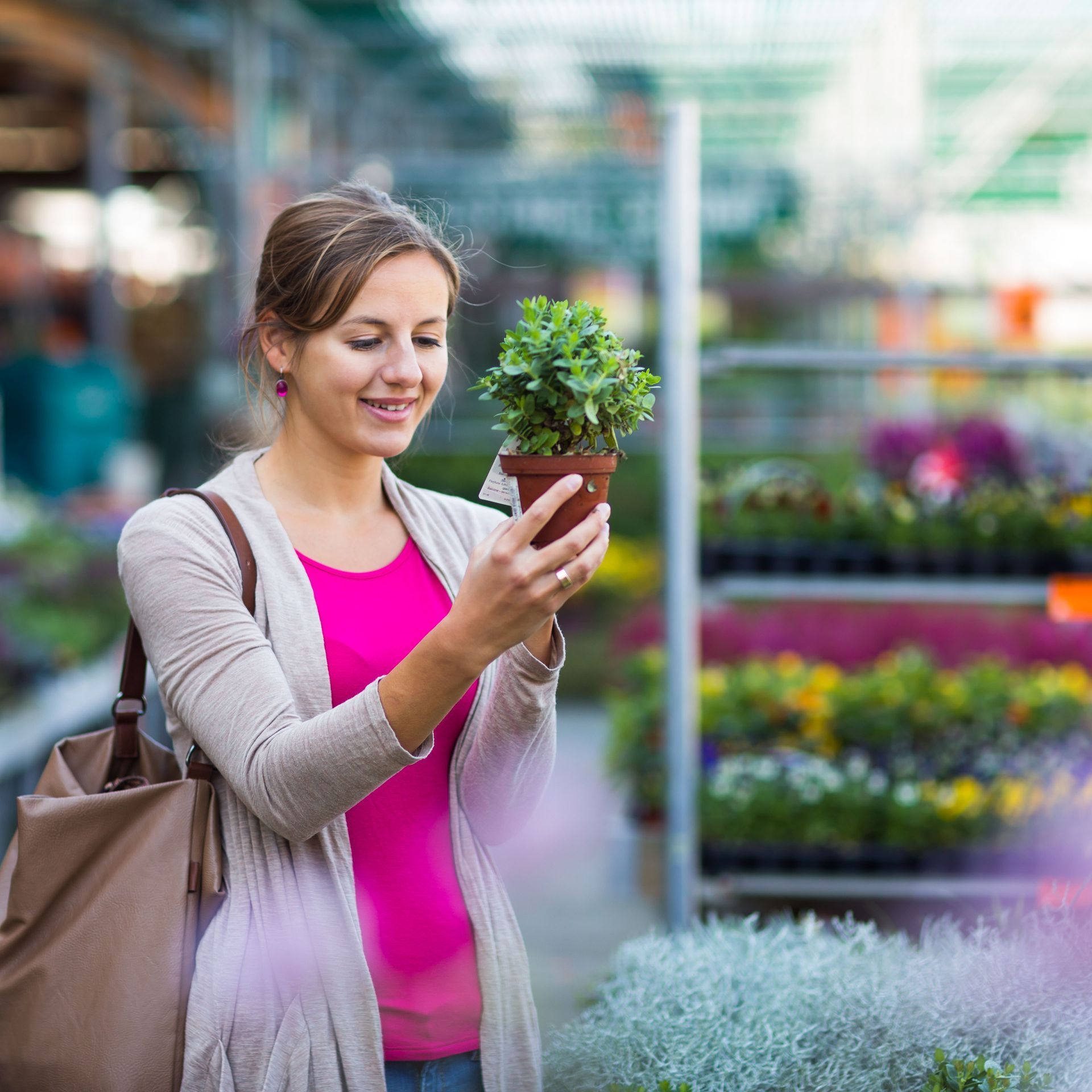 A person in a pink top and beige cardigan smiles while holding a small potted plant in a nursery.