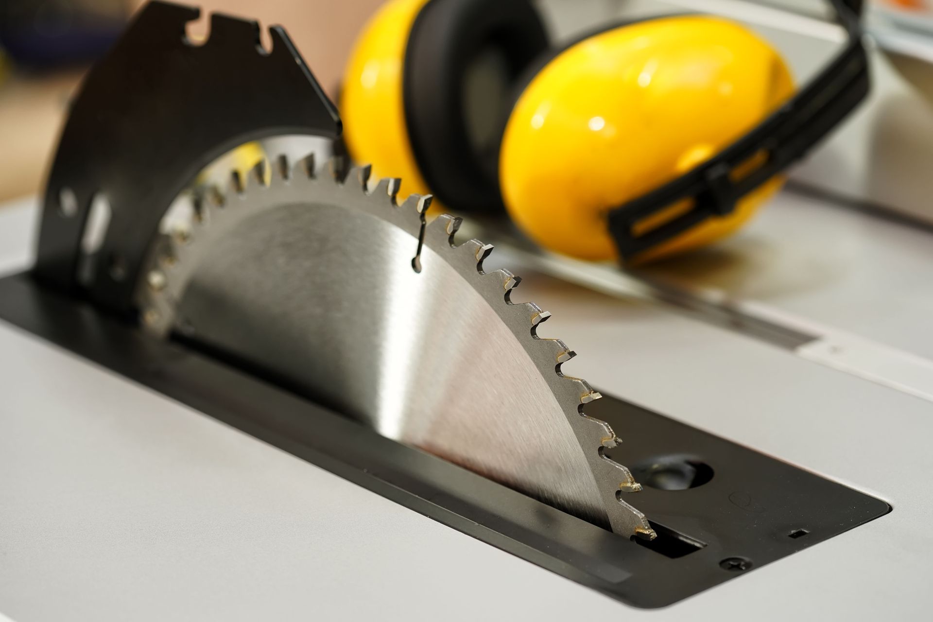 A sharp circular saw blade protruding from a metal table saw, with bright yellow earmuffs resting in the background.