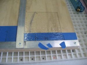 A clear square piece of glass is taped onto a light-colored wooden surface next to an aluminum ruler.