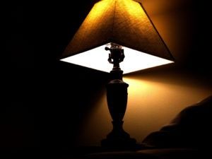 A warm-lit table lamp with a dark shade sits in a dimly lit room, casting a soft glow on a nearby wall.