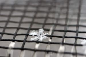 A delicate, intricate snowflake rests on a dark, grid-patterned window screen.