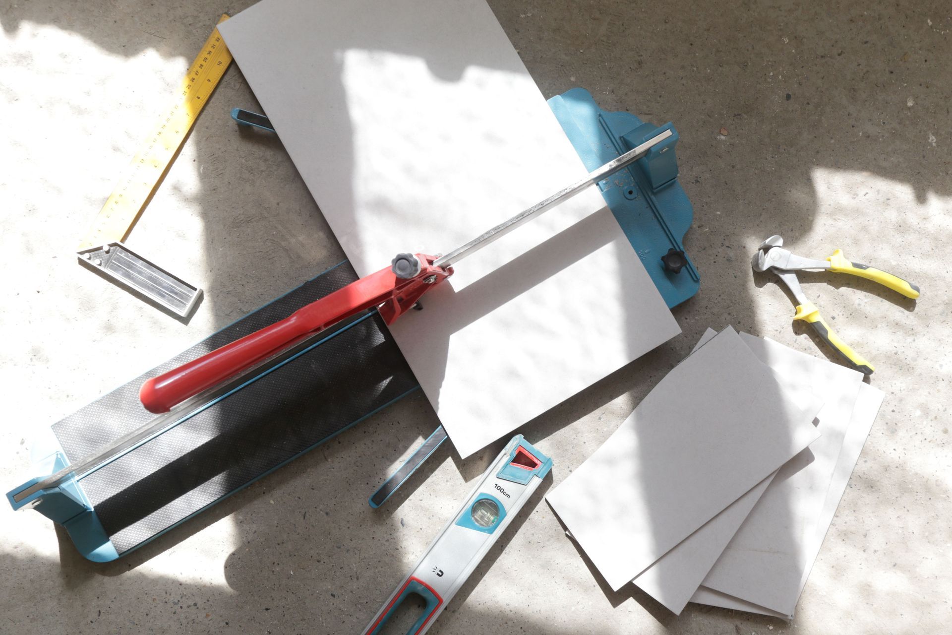 A manual tile cutter with a white tile, a yellow square, tile nippers, and a spirit level on a concrete floor.