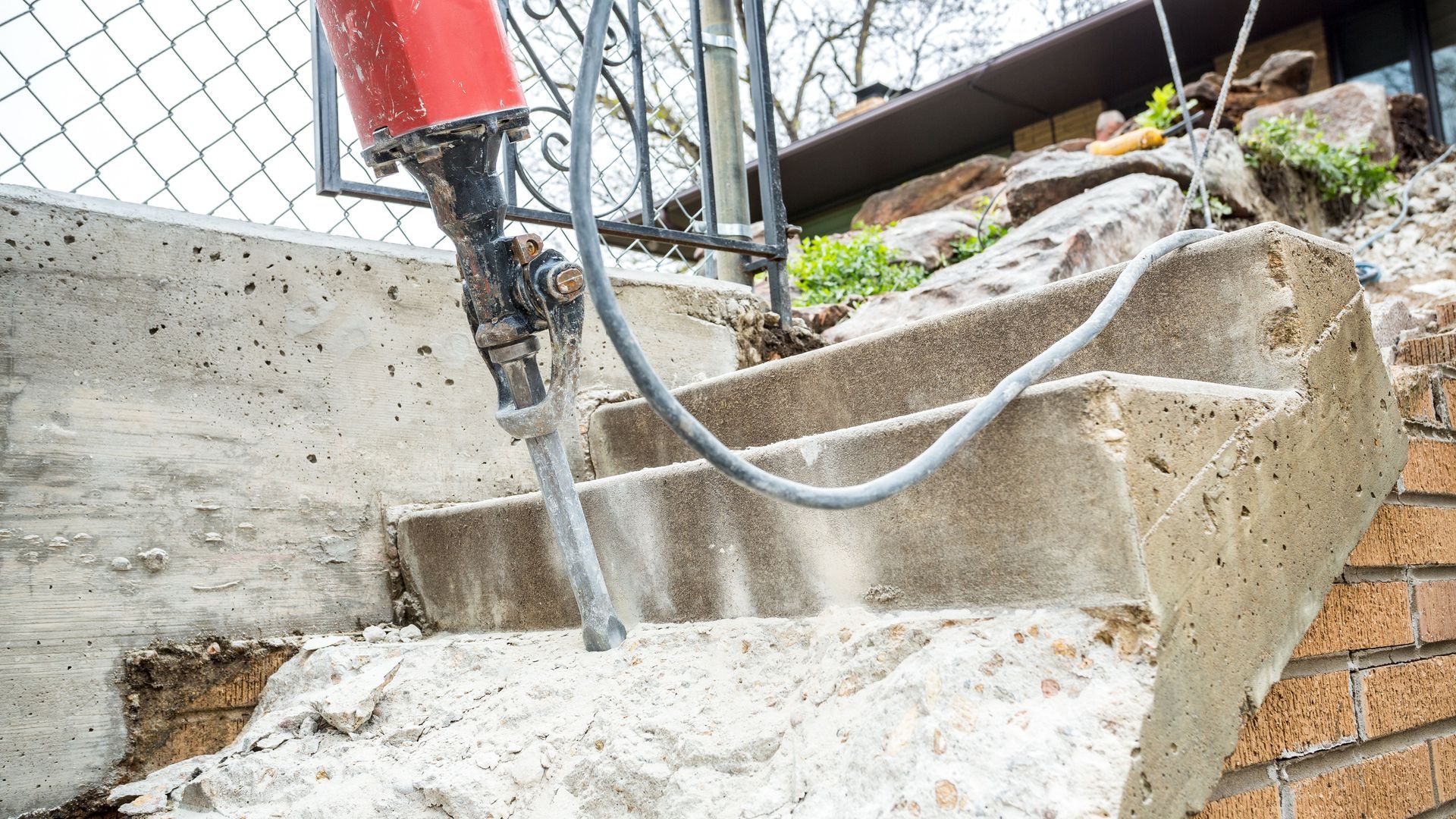 A jackhammer breaking apart concrete outdoor stairs next to a brick wall and a wire fence.