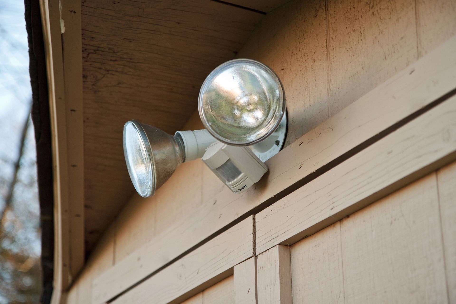 A two-bulb motion-sensor floodlight mounted on the wooden exterior wall of a building.