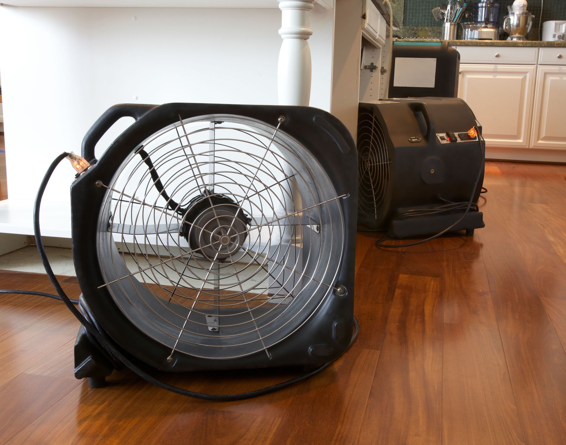 Two black industrial air movers sit on a hardwood floor in a kitchen, used for drying the area.