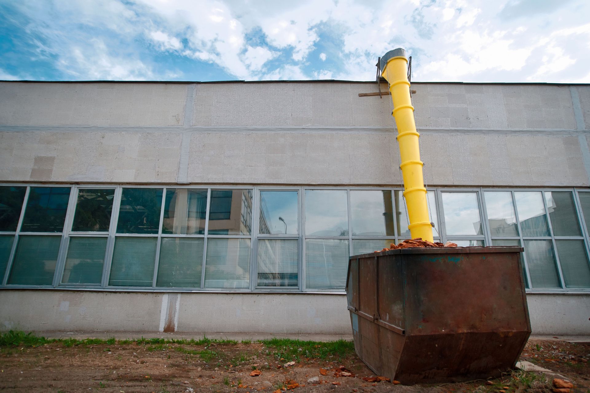 A yellow debris chute extends from the roof of a gray industrial building into a metal dumpster below.