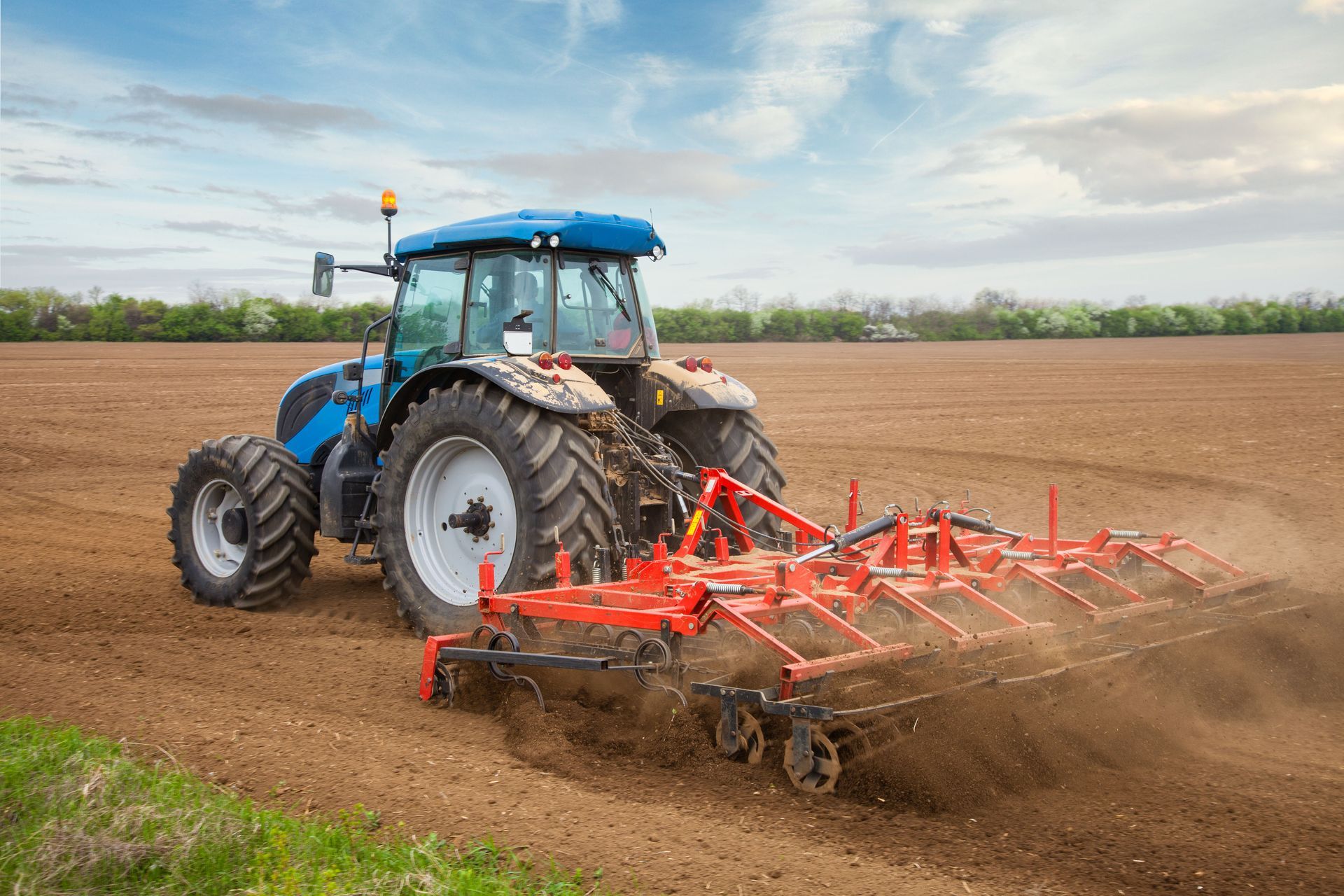 A blue tractor pulls a red cultivator through a brown field under a cloudy sky.