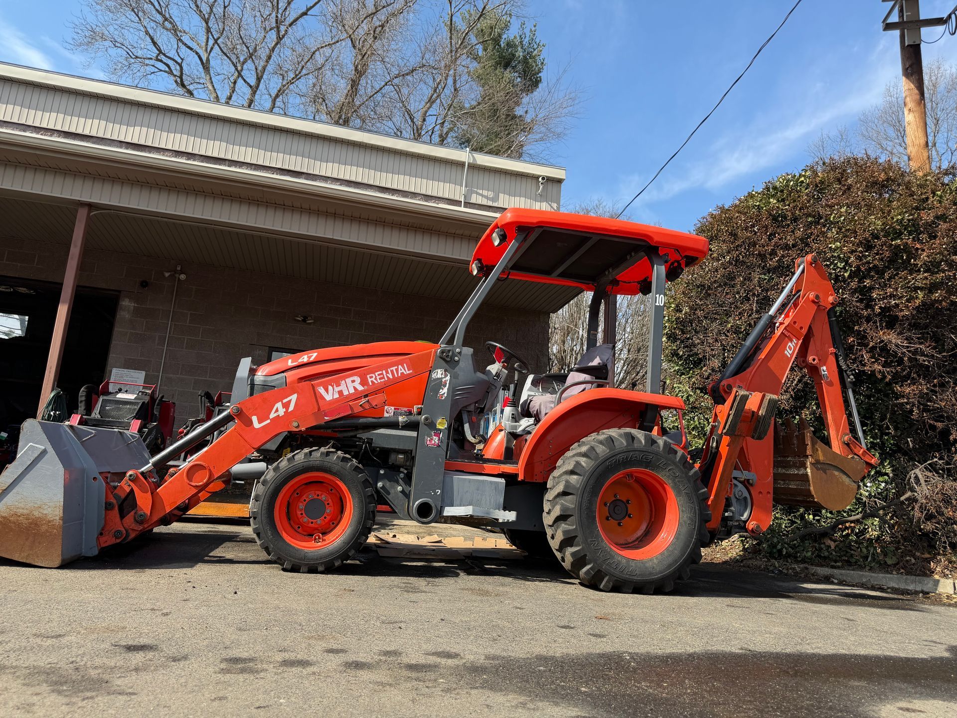 An orange Kubota L47 loader-backhoe parked outdoors on a paved lot next to a brick building.