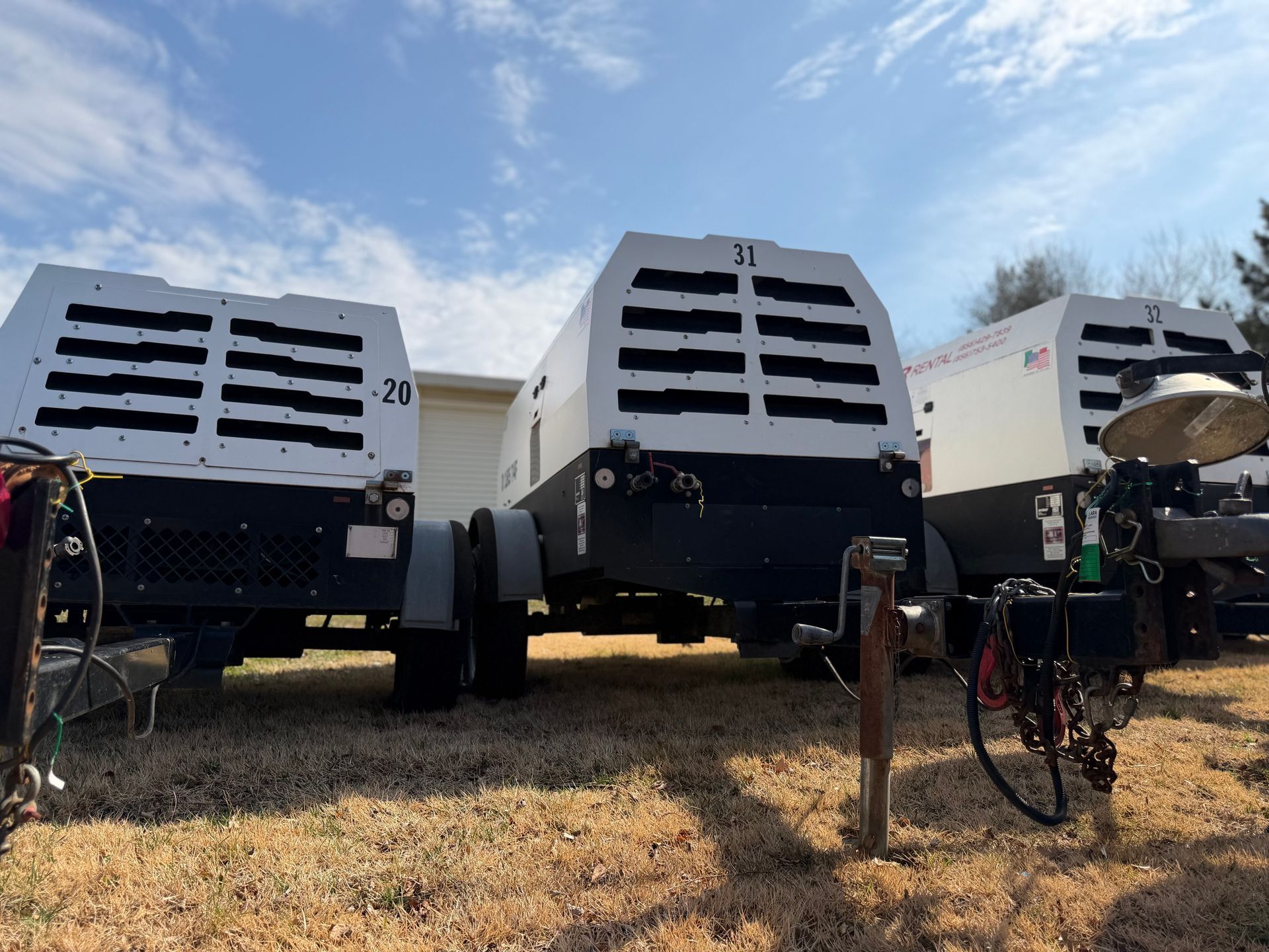 Three white and black portable air compressors parked side-by-side on a gravel lot under a blue, cloudy sky.