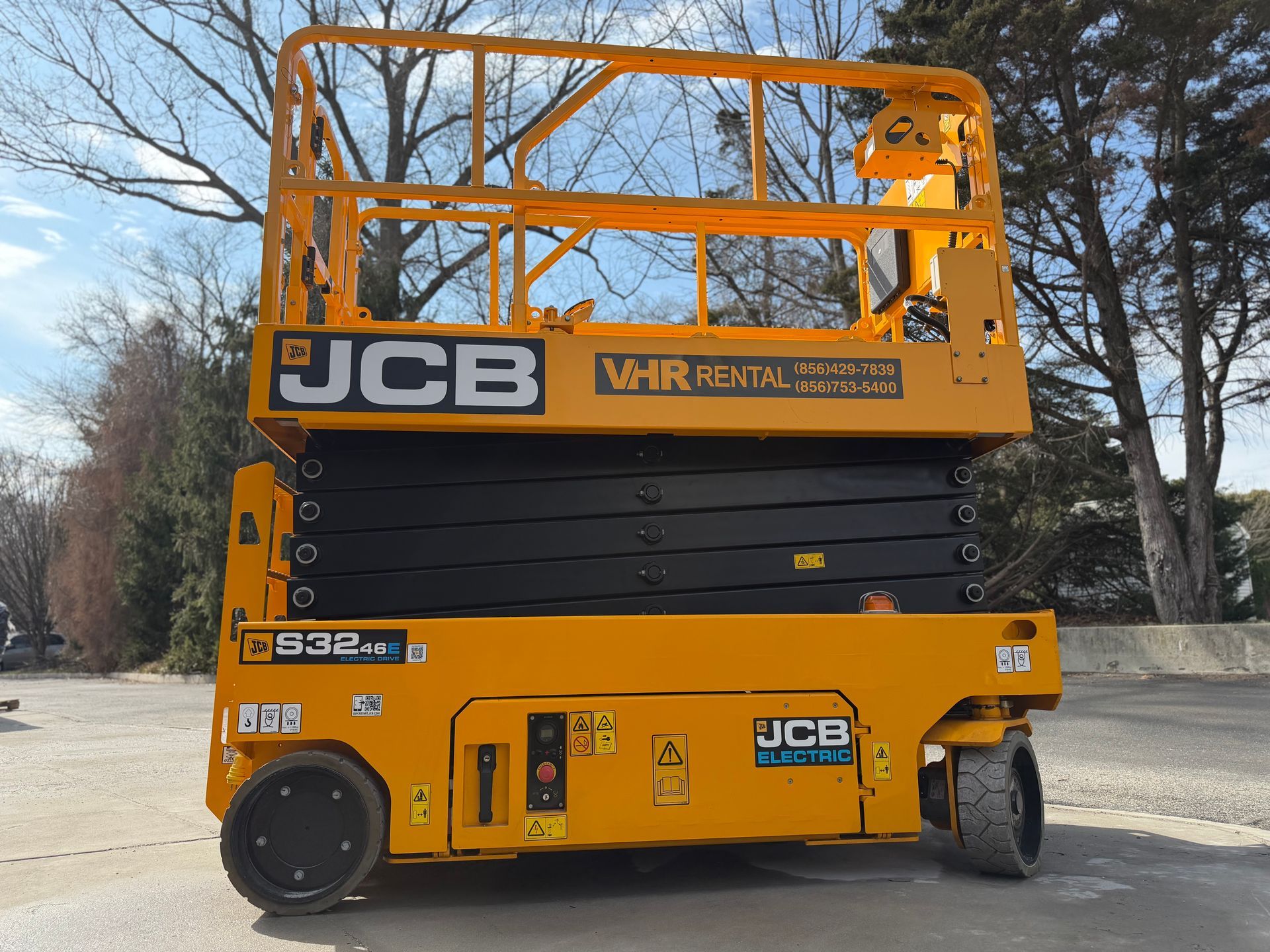 A yellow JCB scissor lift parked outdoors on a gravel lot under a clear, sunny sky.