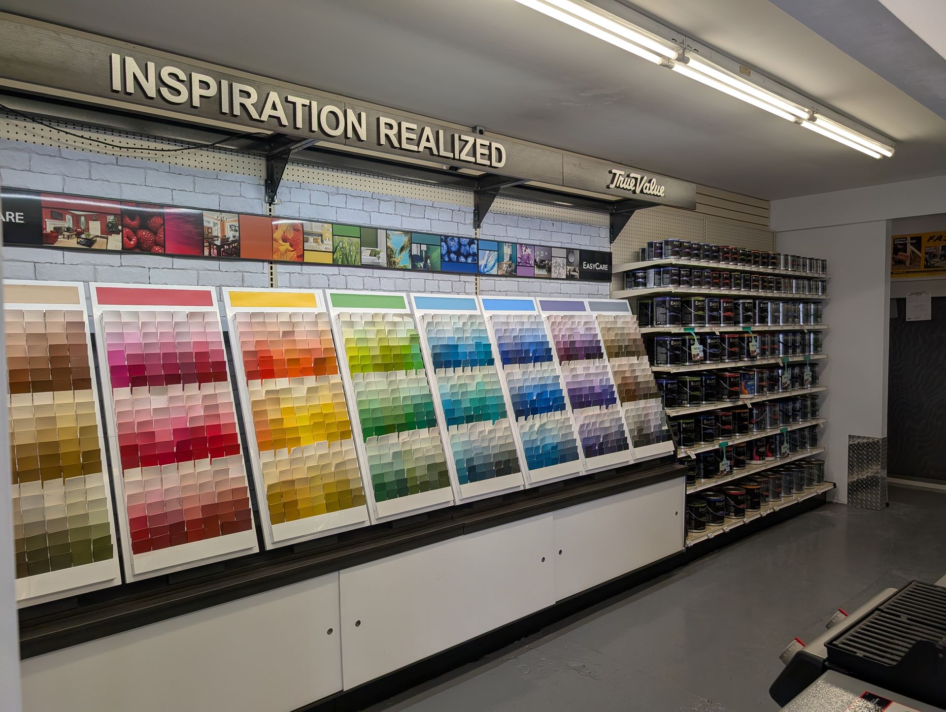 A paint store aisle featuring a large display of color sample swatches arranged by hue with paint cans on adjacent shelves.