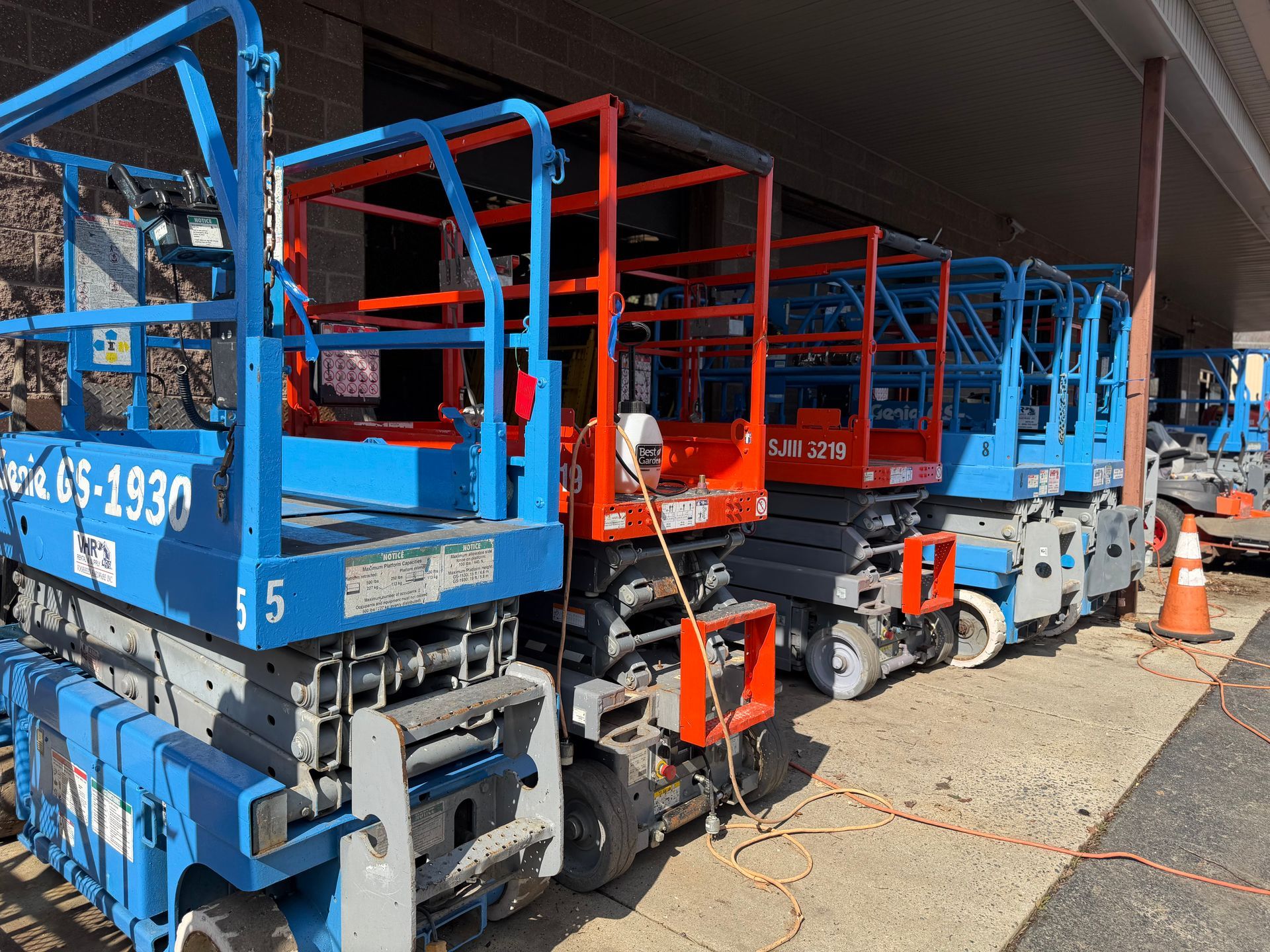 A line of several blue and red scissor lifts parked in a row outdoors under a building overhang.