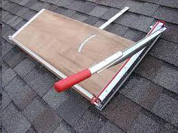 A portable roofing shingle cutter with a wooden base and a red-handled metal lever, resting on a shingled roof.
