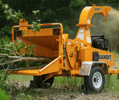 A bright yellow Bandit wood chipper processes tree branches in a grassy, wooded area.