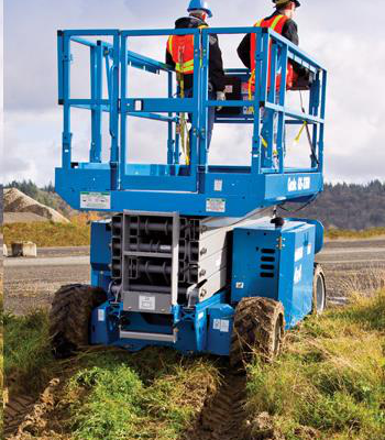 Two workers in safety gear operate a blue scissor lift on a dirt and grass job site.
