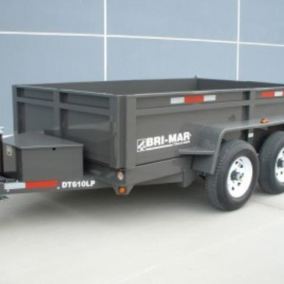 A gray Bri-Mar DT610LP dump trailer sits on a paved surface in front of a blue paneled wall.