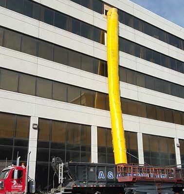 A yellow construction chute extends from an upper-floor window of a white office building into a dumpster on a truck.