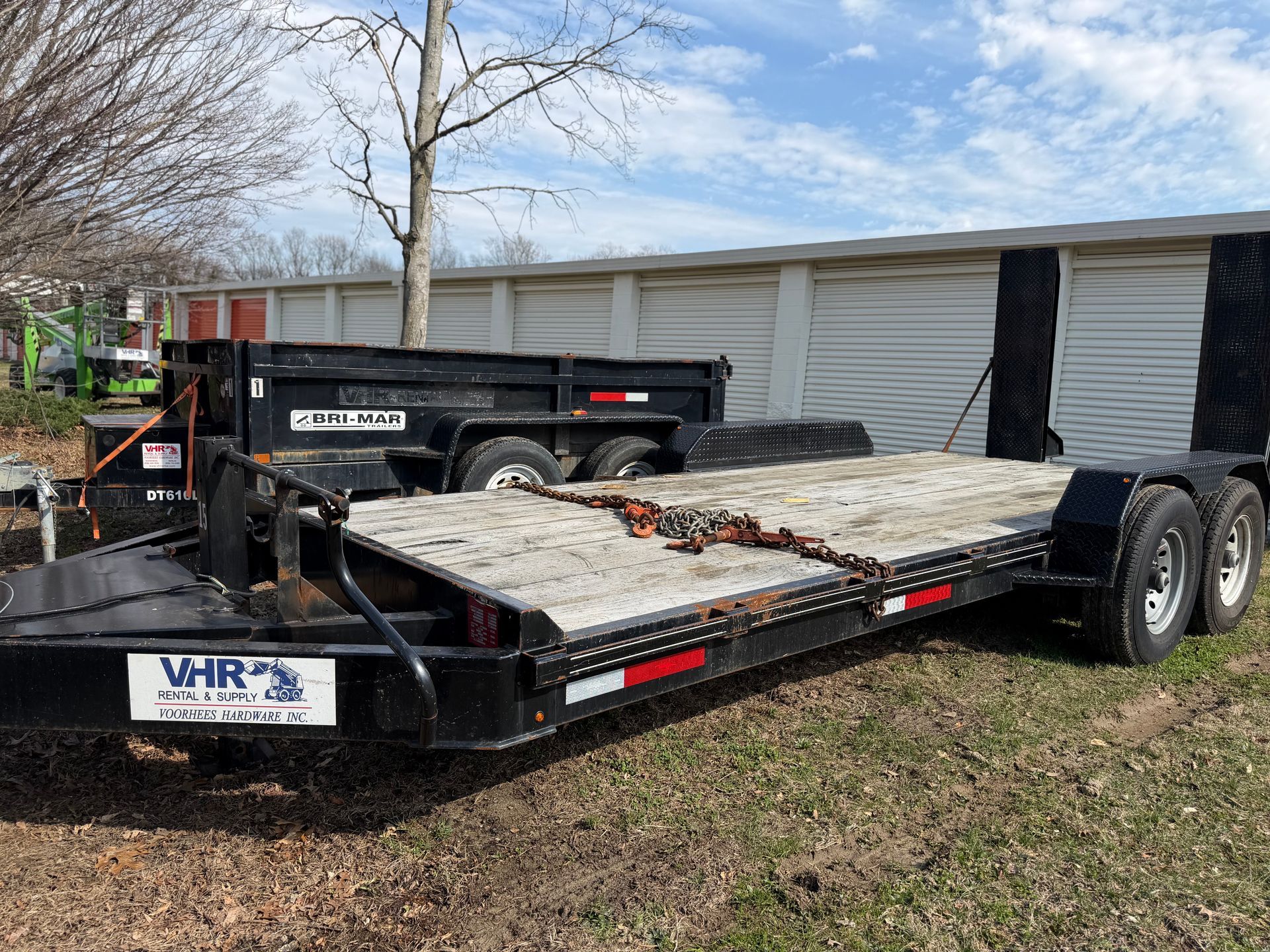 A black flatbed equipment trailer parked on grass in front of a storage facility.