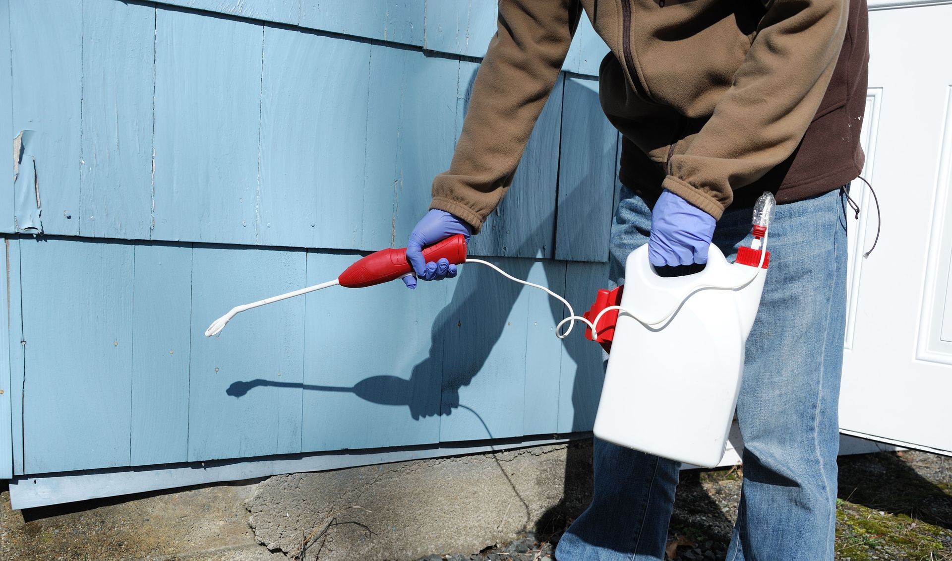 A person in a brown jacket and gloves uses a handheld pump sprayer to treat the exterior of a light blue house.