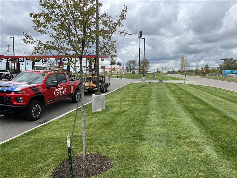 A red work truck with a trailer parked next to a large, freshly mowed grassy lawn on a cloudy day.