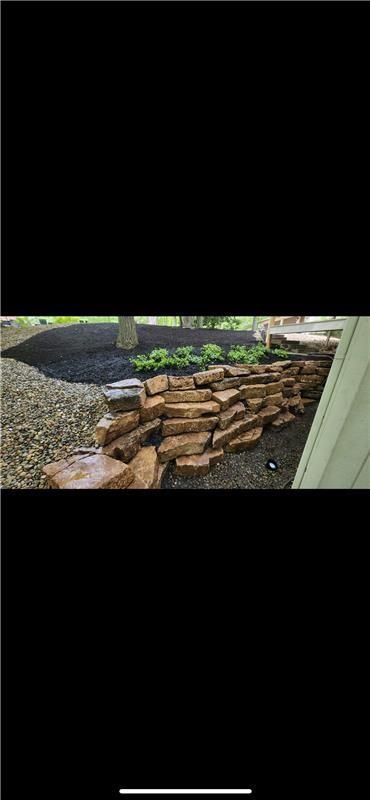 Stone retaining wall with plants and mulch, beside a house, in a garden.