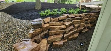 A low, stacked-stone retaining wall curves along a landscaped yard with dark mulch, small green plants, and gravel.