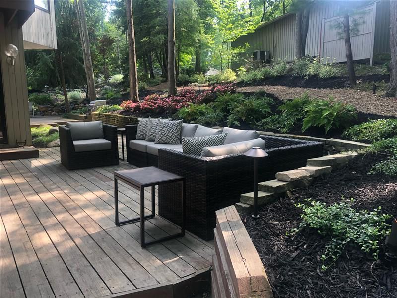 A dark wicker sectional and armchair with light gray cushions sit on a wooden deck next to a landscaped garden.