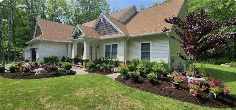 A light-colored house with a brown roof, surrounded by mulched garden beds and greenery on a sunny day.