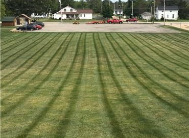 A wide expanse of lawn with evenly mown grass stripes leading toward a parking lot and houses in the background.