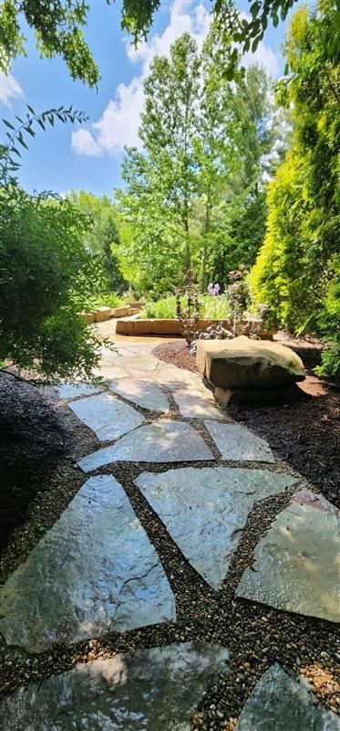 Stone path winds through a lush garden under a blue sky, surrounded by trees and greenery.