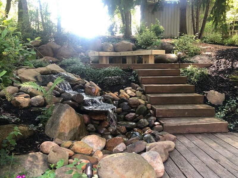 A stone waterfall flows beside wooden stairs leading to a bench in a lush, landscaped garden.