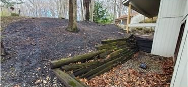 A low, weathered timber retaining wall sits next to a house near a sloped, dark, mulch-covered yard and bare trees.