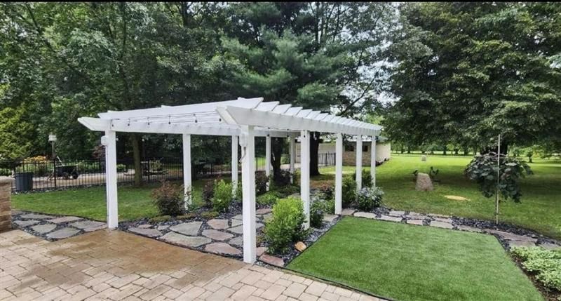 White pergola over stone path and green lawn in a backyard, surrounded by trees and bushes.