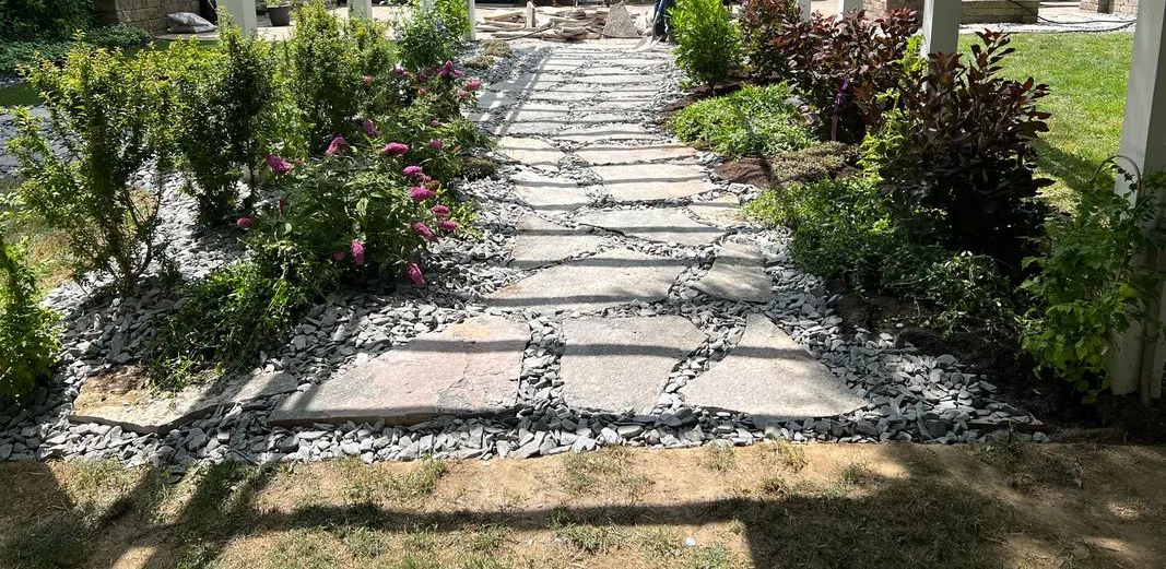 Stone path through garden under white pergola; two people working in the background.
