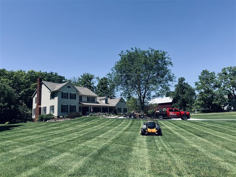 A lawn mower on a large green lawn in front of a house, with a red truck parked near a barn in the background.