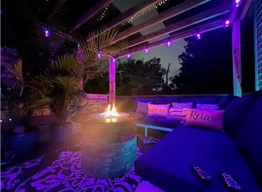 A cozy outdoor lounge with a fire pit, plush blue seating, purple string lights, and patterned rug at twilight.
