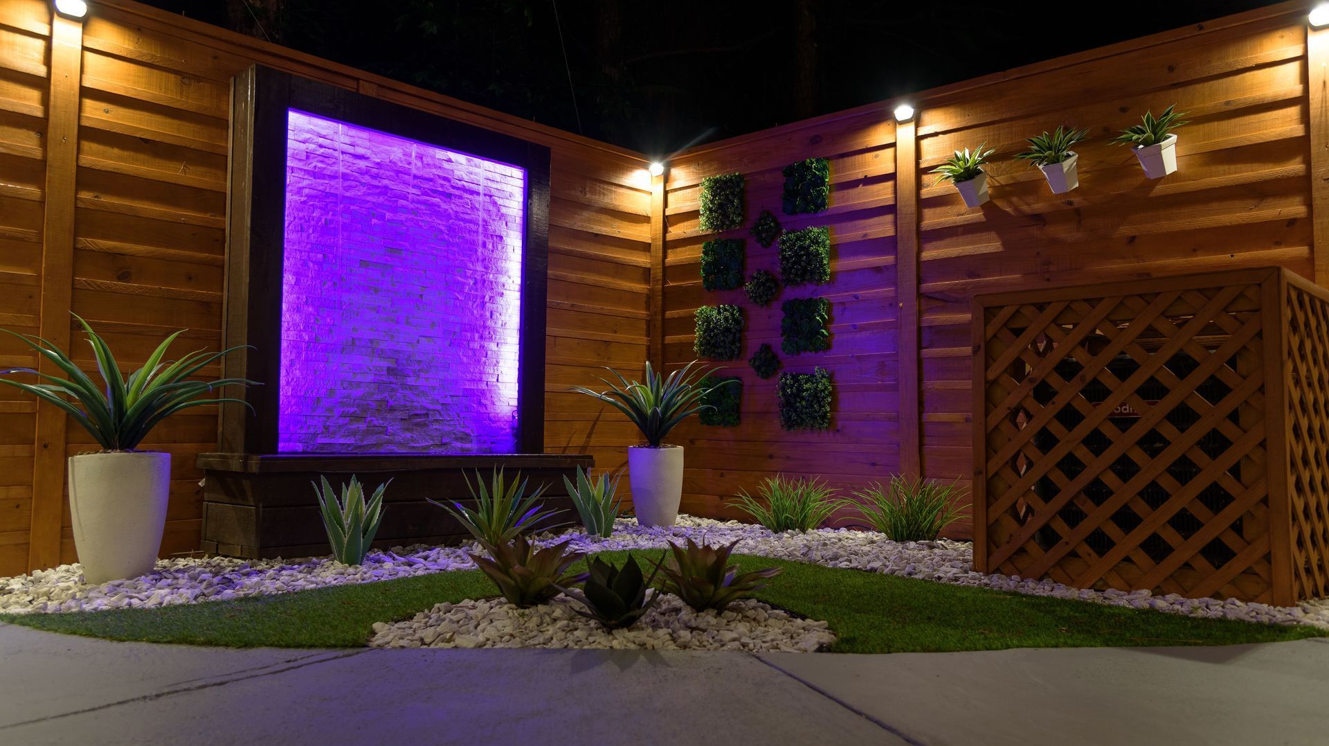 A backyard patio at night features a glowing purple waterfall, wooden privacy fence, potted plants, and landscape lighting.