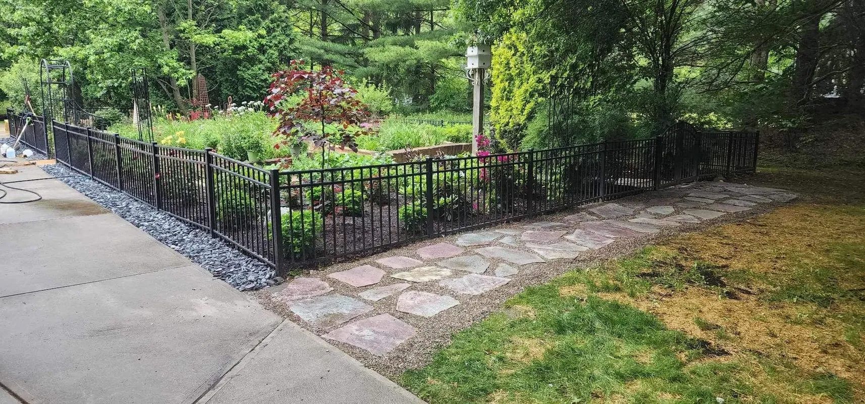 Black metal fence encloses a garden bed with a stone walkway. Green grass and trees are in the background.