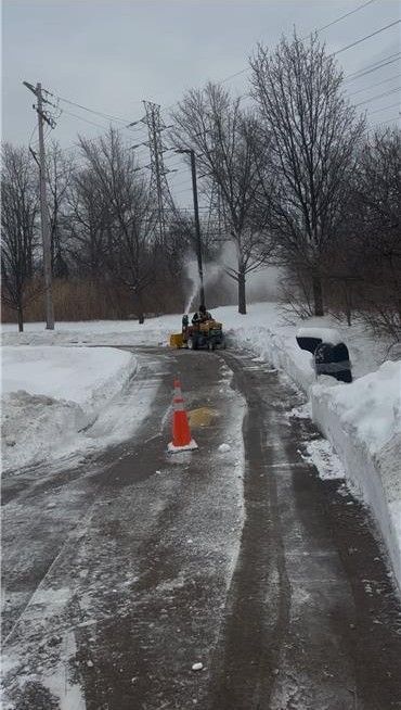 Snow blower clearing a snowy road with a cone, power lines and trees.