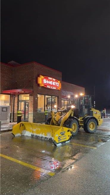 A yellow snow plow sits in a parking lot in front of a brightly lit Sheetz store at night.