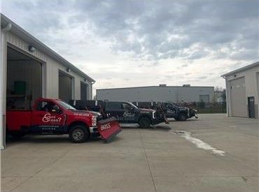 Red trucks with snowplows parked in front of a building with open garage bays on a cloudy day.