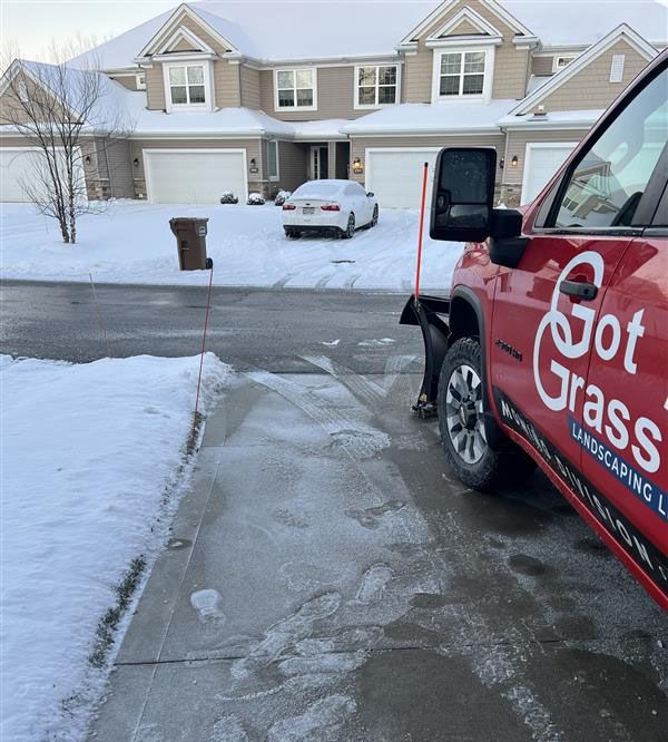 A red landscaping truck with a snowplow sits on a snowy driveway in front of a multi-unit house on a winter day.