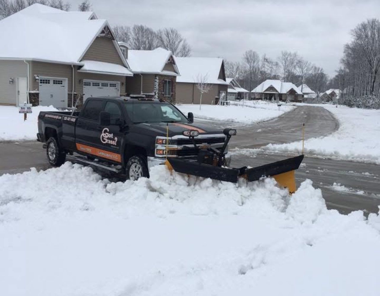 A dark-colored pickup truck with a plow attached clearing snow from a residential street on a cloudy winter day.