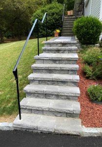 A set of gray stone outdoor stairs leading up to a house, featuring a black metal handrail on the left side.