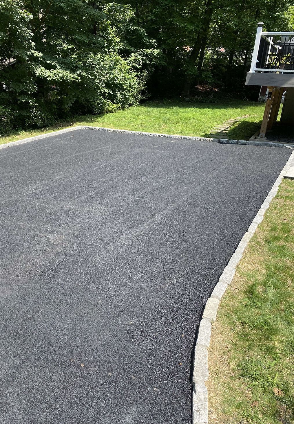 A newly paved dark asphalt driveway with stone border edging, situated next to a grassy yard and a deck.